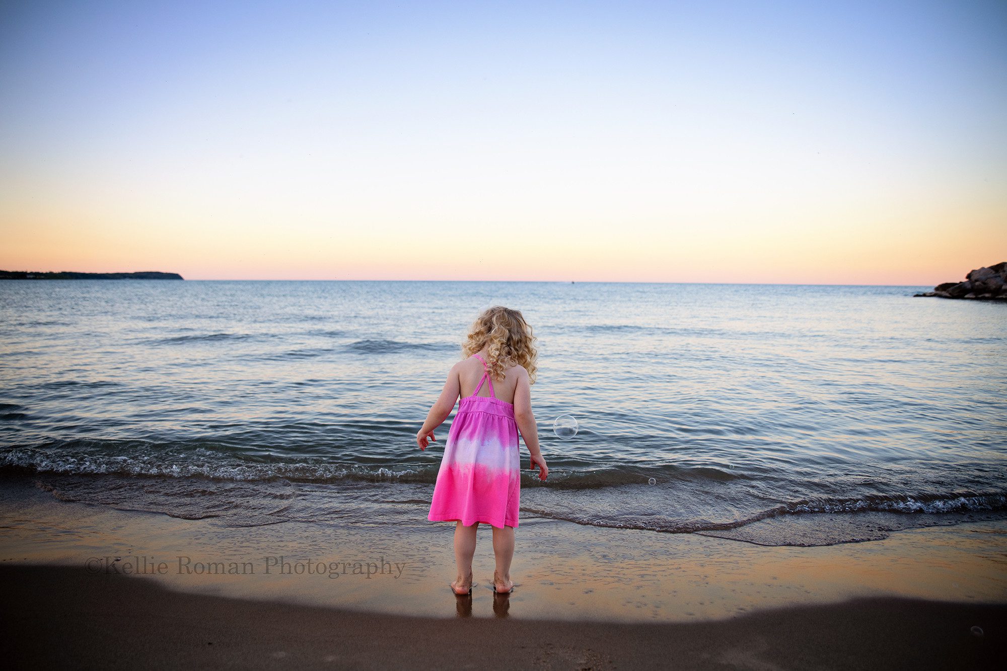 bubbles at the beach a three year old girl standing on a beach in milwaukee looking down at the water she has on a bright pink dress and has long curly blonde hair the sky and water are lit up with shades of pastel during sunset