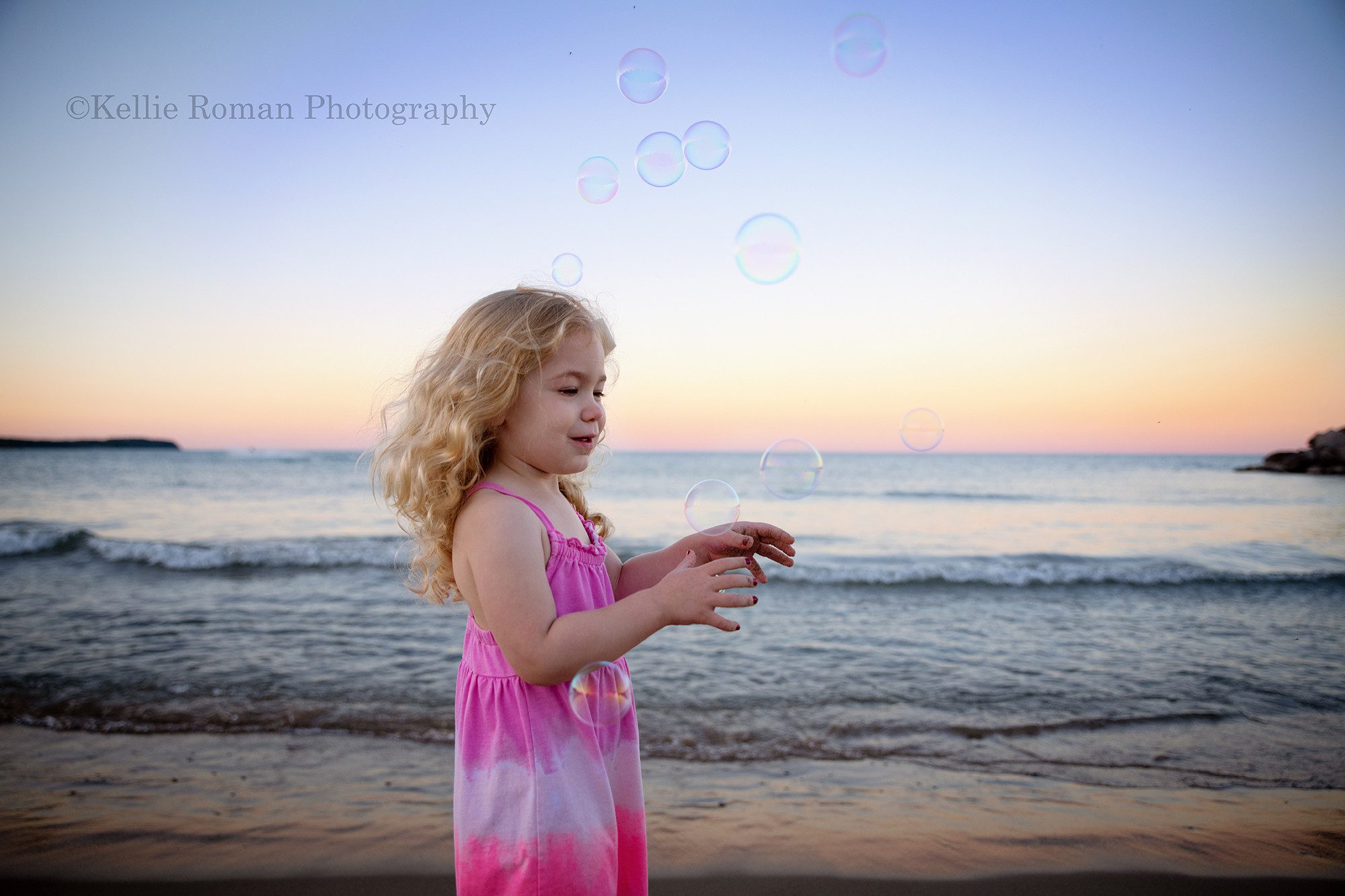 bubbles at the beach a three year old girl on a milwaukee beach wearing a pink dress is playing with bubbles floating through the air she has long curly blonde hair and the sky behind her is lit up with shades of pastel during sunset