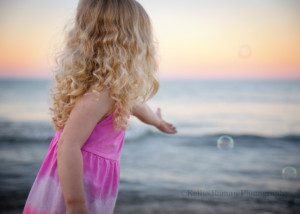 bubbles at the beach a toddler girl wearing a pink dress is holding her hand out for several colored bubbles floating through the air its a shot of the back of her head she has long curly blonde hair being photographed on a milwaukee Wisconsin beach the sunset has lit up the sky with shades of pastel