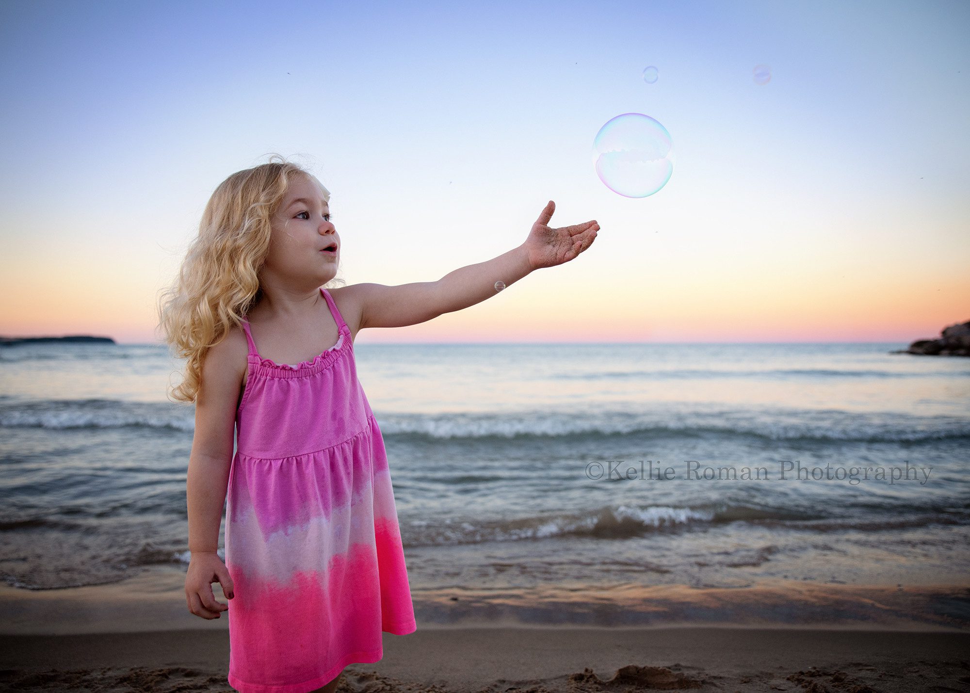 bubbles at the beach a three year old girl with blonde curly hair is standing on the beach with her arm up in the air ready to catch a bubble she is wearing a pink dress and the sunset sky is very colorful behind her