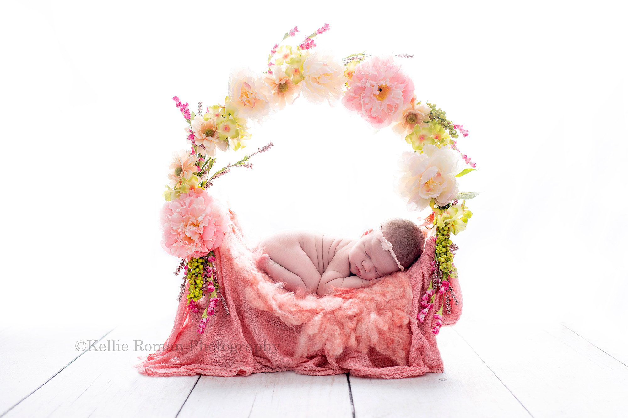 baby photos a newborn baby girl is curled up on her tummy on a floral ring bed the bed has pink and green flowers surrounding her light a wreath the bottom has different textures of pink fabric the baby is sleeping and has a small pink bow headband on there is backlighting so the image is very bright and white