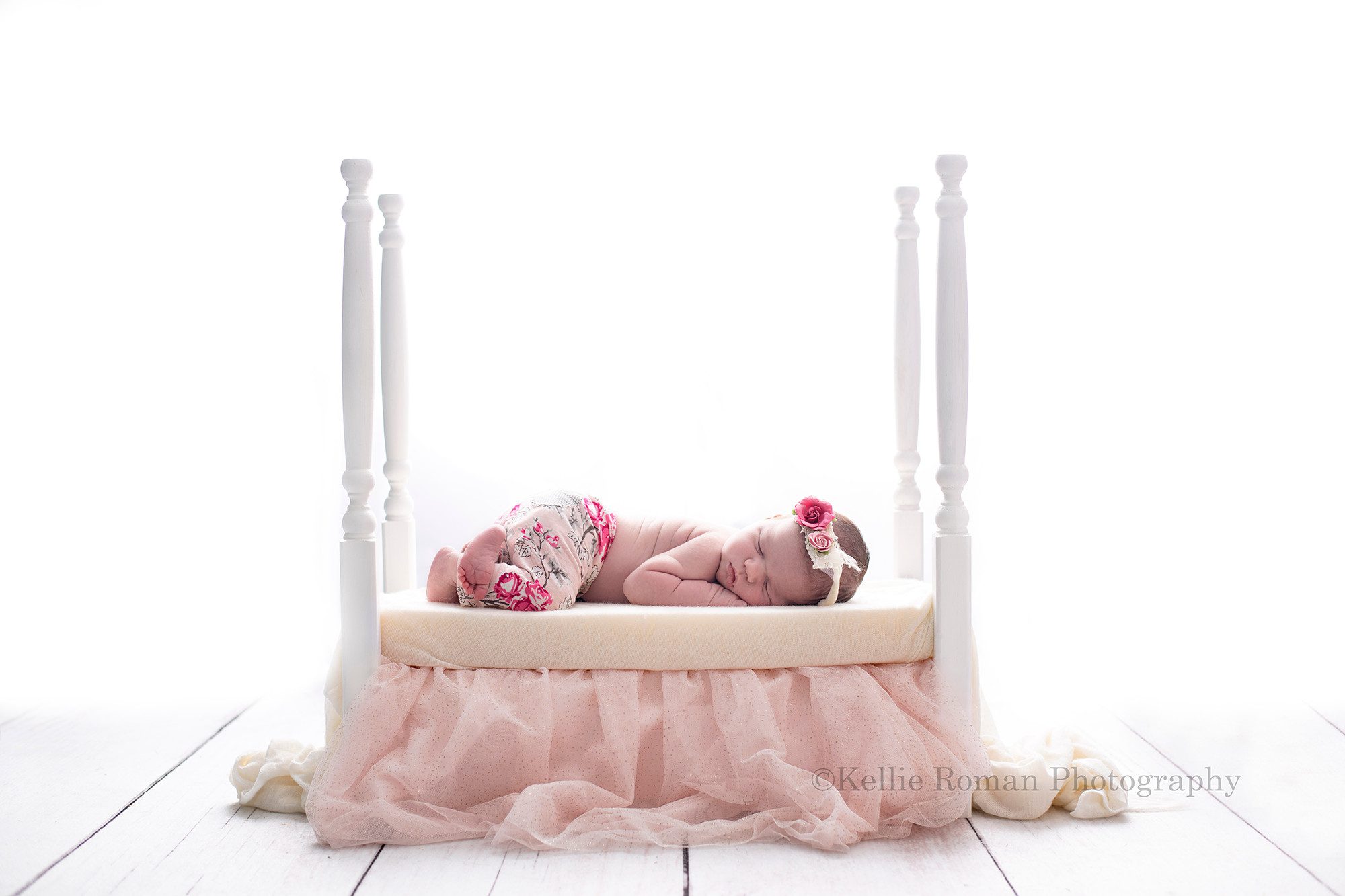 baby photos a newborn baby girl laying on her belly onto of a white wood bed with 4 tall bed posts the baby has floral pink pants on and a pink floral headband the bed has a light pink bedskirt and a fabric covering it there is backlighting so the image is very bright and white