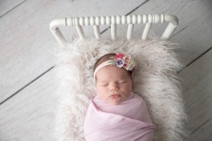 baby photos a infant baby girl in milwaukee Wisconsin photo studio she is wrapped in a light purple fabric and is sleeping she is laying on a white bed that has cream fur onto of it she is wearing a flower headband with shades of pink and purple