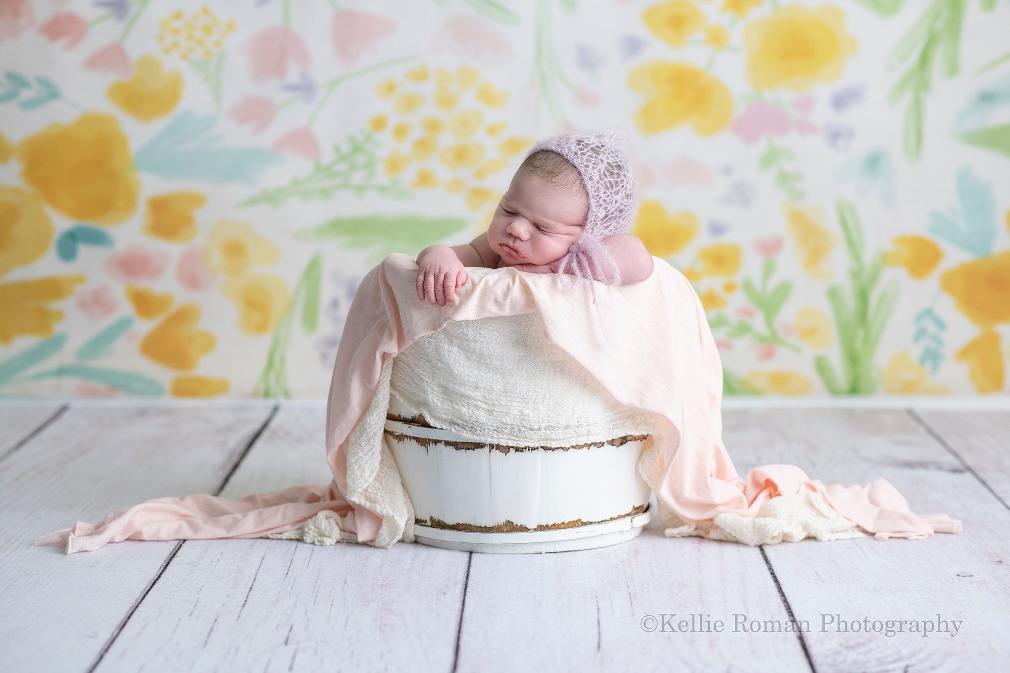 baby photos a newborn infant girl in a milwaukee Wisconsin photographer studio she is posed upright in a white wood bucket the bucket is onto of a white wood floor and in front of a floral backdrop with shades of yellow pink purple and blues. The newborn is resting her chin on one of her arms and the other arm is over the edge of the bucket she is wearing a pink bonnet