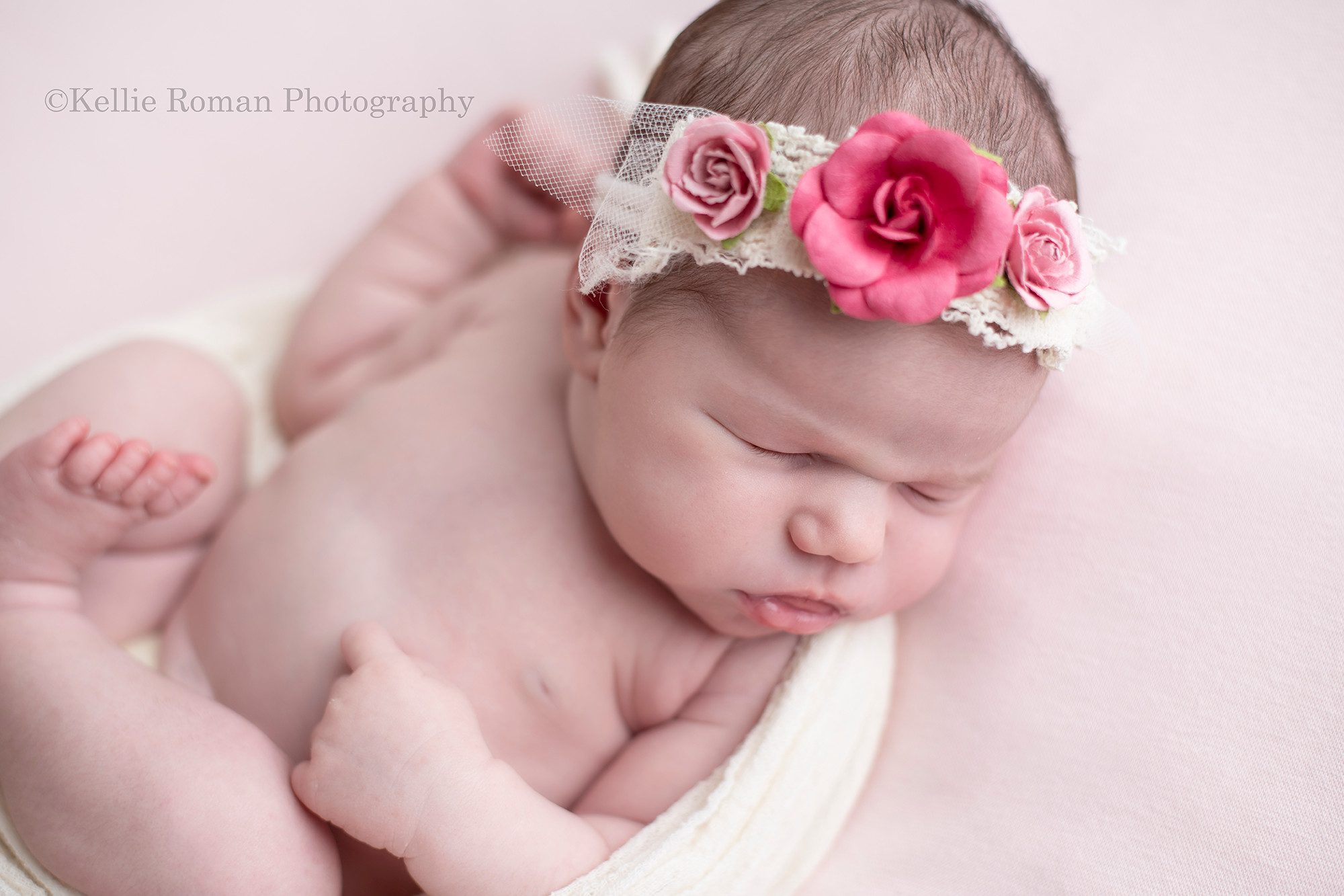 baby photos a little newborn girl in a milwaukee photo studio sleeping on top of a pink blanket posed on a bean bag she is sleeping on her back with a cream swaddle wrap around her she has her hands resting on her tummy and is wearing a pink and purple flower headband
