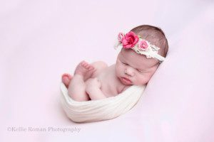 baby photos a newborn little girl in a milwaukee photo studio she is wrapped like an egg with a cream colored wrap she's sleeping on her back with her hands resting on her chest her feet are curled into her chest and she is wearing a flower headband