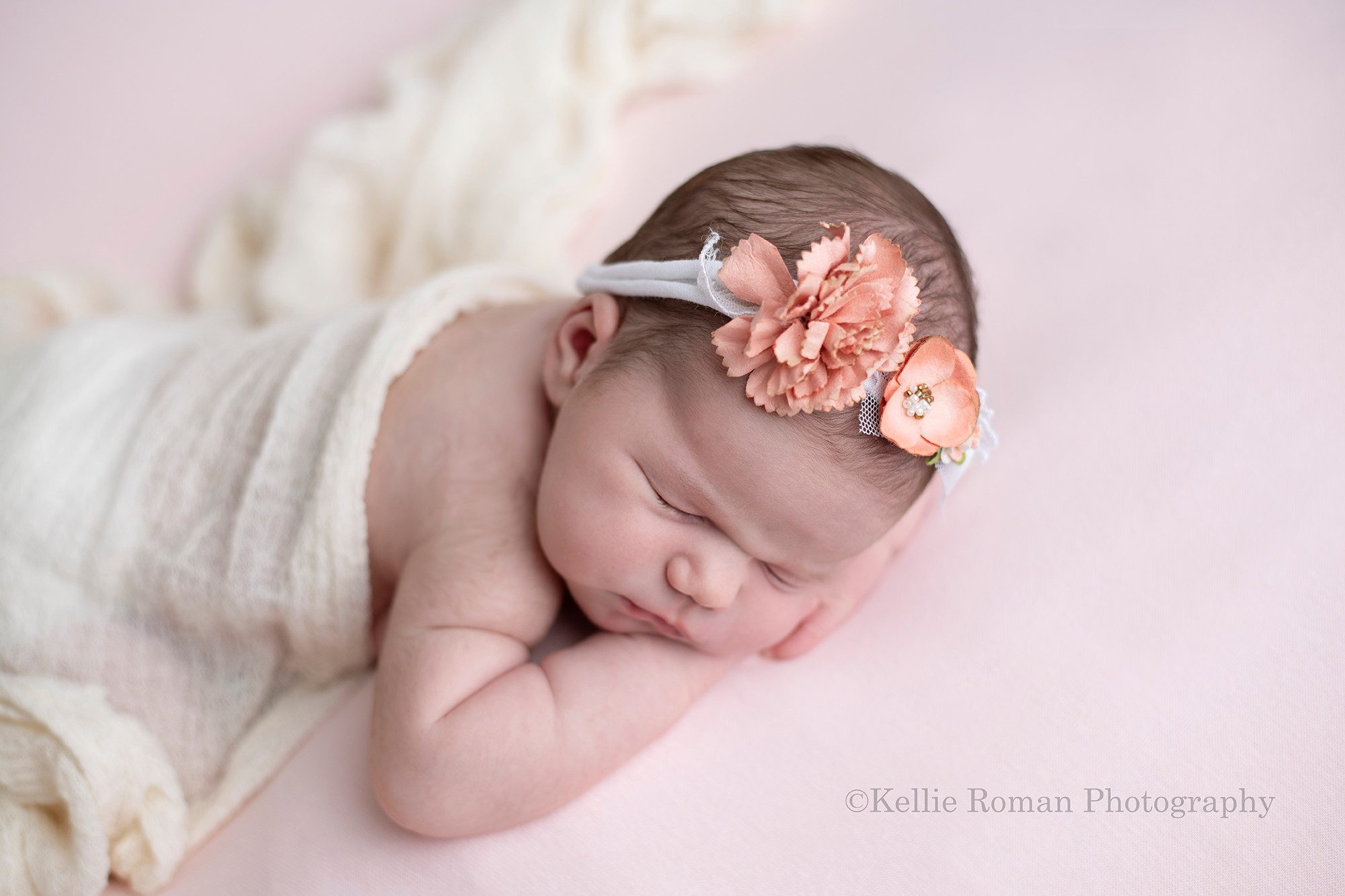 baby photos a newborn baby girl posed onto of a light pink blanket in milwaukee photo studio she is sleeping on her belly with her chin resting on her hands she is covered in a cream fabric and is wearing a floral headband