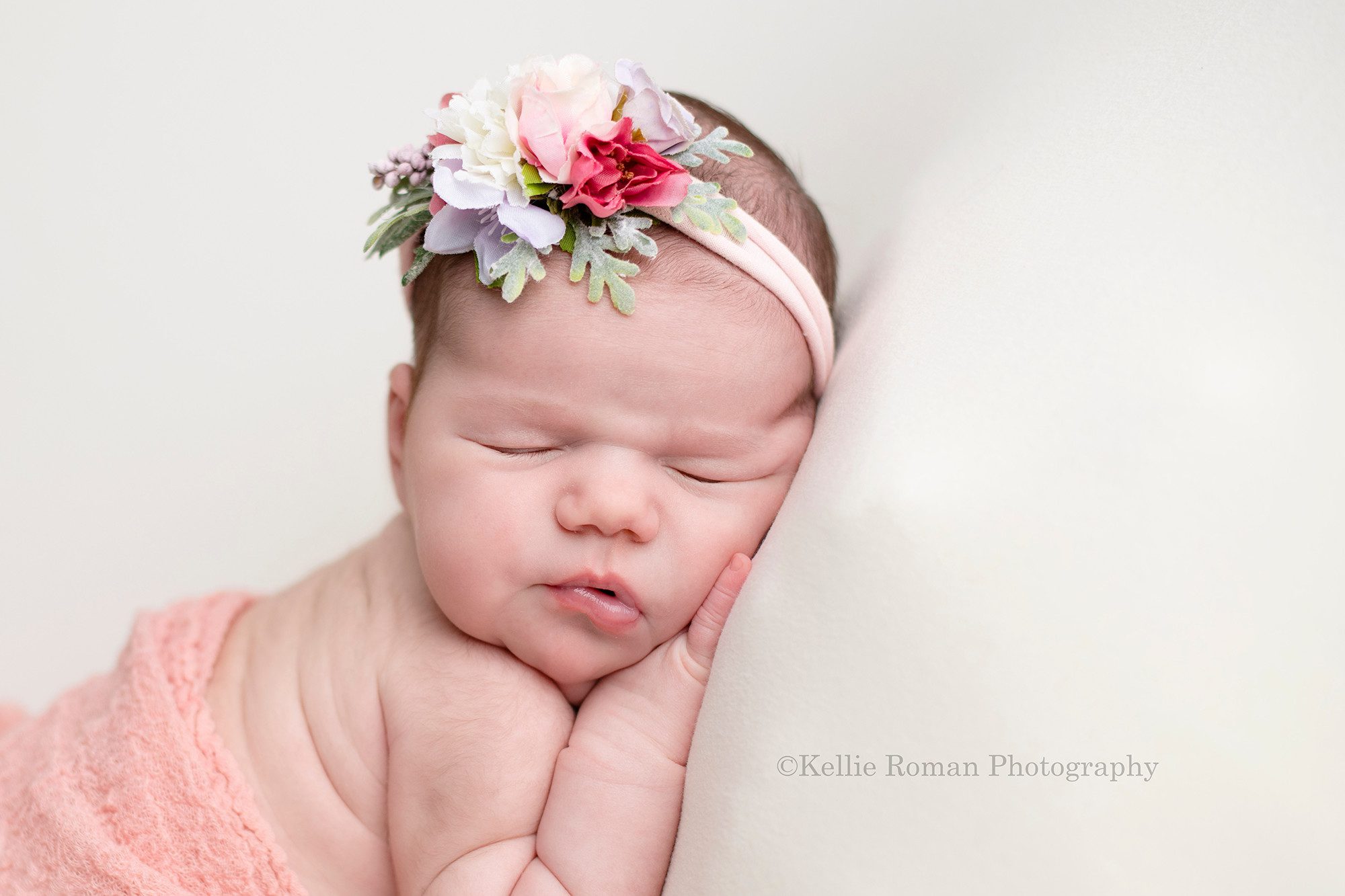 baby photos in milwaukee photographer studio a baby girl posed with her hand under her chin on top of a cream colored blanket she has a floral pink and purple headband on and is sleeping on her belly