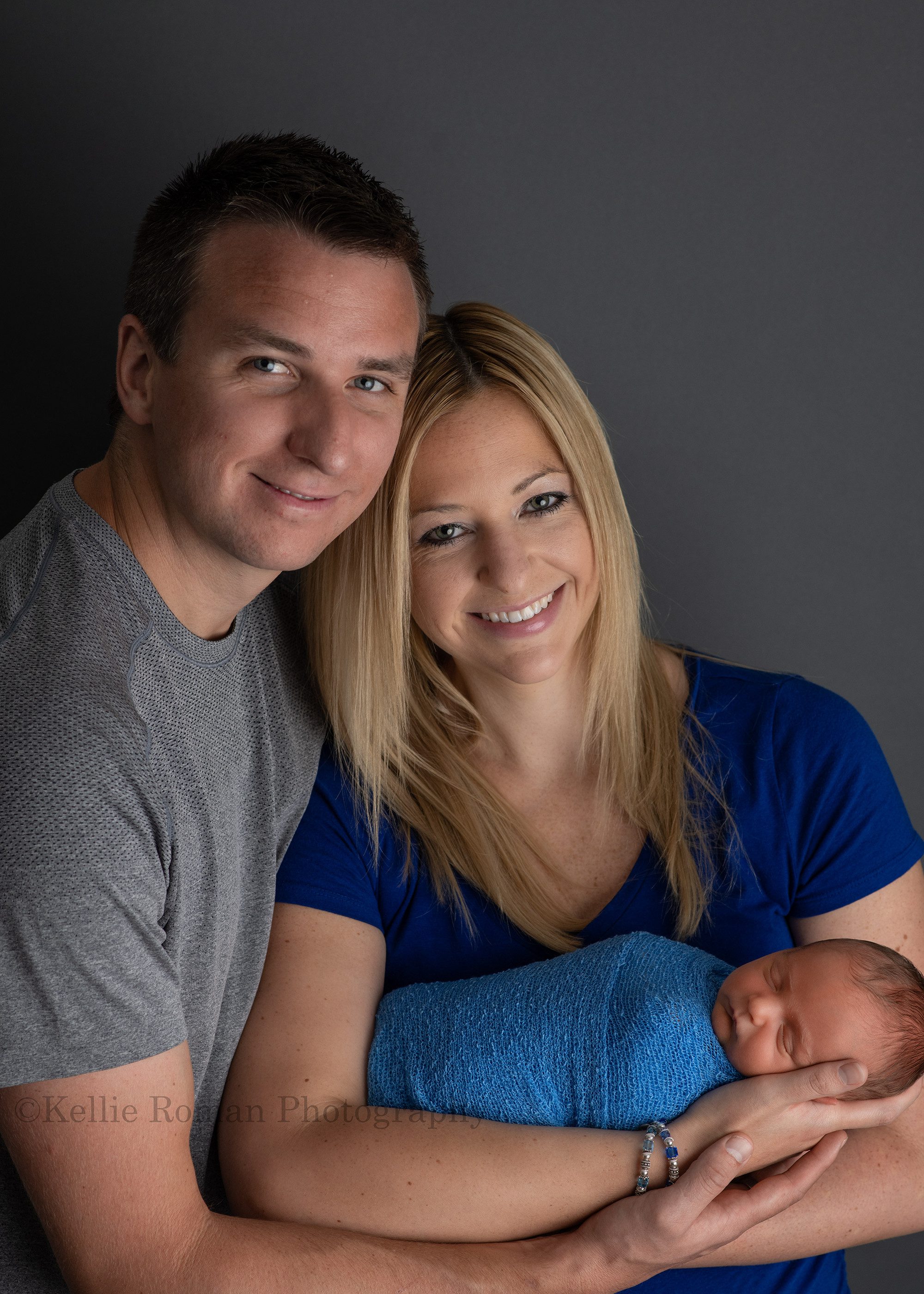 rainbow baby a shot of mother and father holding onto newborn baby boy who is wrapped in a blue swaddle the couple is wearing blue and grey and is looking up at the camera the image is taken in milwaukee photographers studio