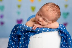 rainbow baby a close up image of a newborn boy in a white wood bucket he is posed upright with his chin on his hands and he is on top of a blue polka dot fabric the backdrop behind him is blue with rainbow colored hearts in a Milwaukee photographers studio