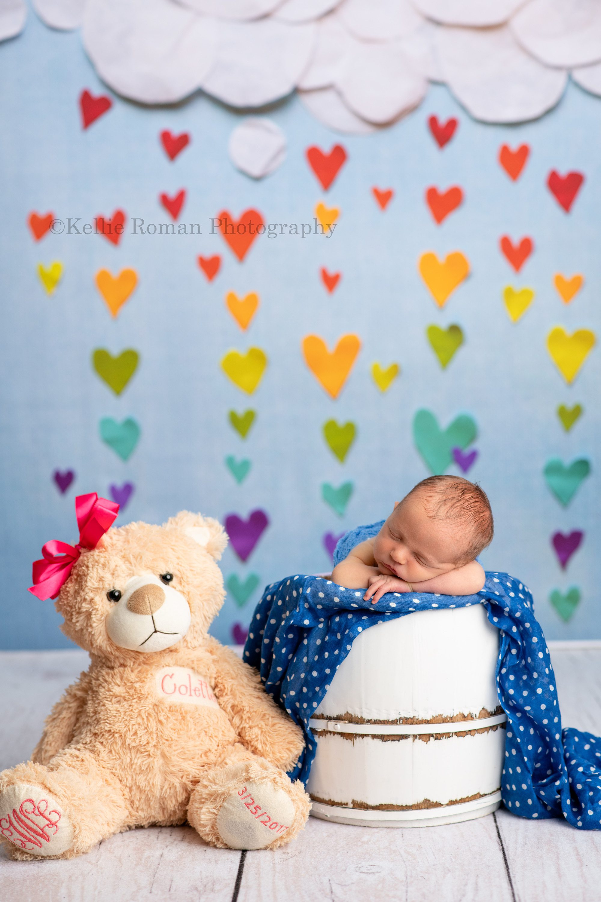 rainbow baby a newborn boy is posed in a white bucket in front of a blue backdrop with white clouds and rainbow colored hearts he has his chin on top of his hands and is on top of a blue polka dot fabric next to him on the floor is a brown and pink teddy bear representing his big sister who past away