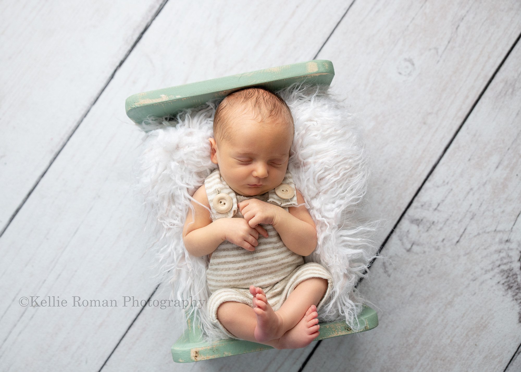 rainbow baby a newborn infant boy sleeping on his back on top of cream colored fur which is placed in a mint colored wood bed the bed is on top of white wood flooring the baby boy is wearing a cream and brown striped romper with big wood buttons he's being photographed in milwaukee photo studio