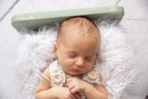 rainbow baby a baby newborn boy in a cream and brown striped romper with big wood buttons is laying on his back onto of cream fur the fur is placed in a teal wood bed the baby Is sleeping and has his hands on his chest being photographed in milwaukee photo studio