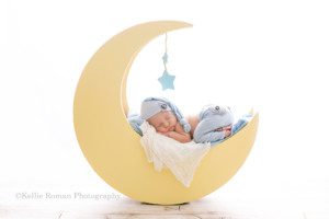 rainbow baby a newborn baby boy posed on his stomach sleeping on top of a yellow moon made out of wood he has his hands under his chin and there are two blue wood starts hanging above him he has on light blue pants and a match sleepy hat he is laying on top of white and blue fabric and the image is very brightly lit up