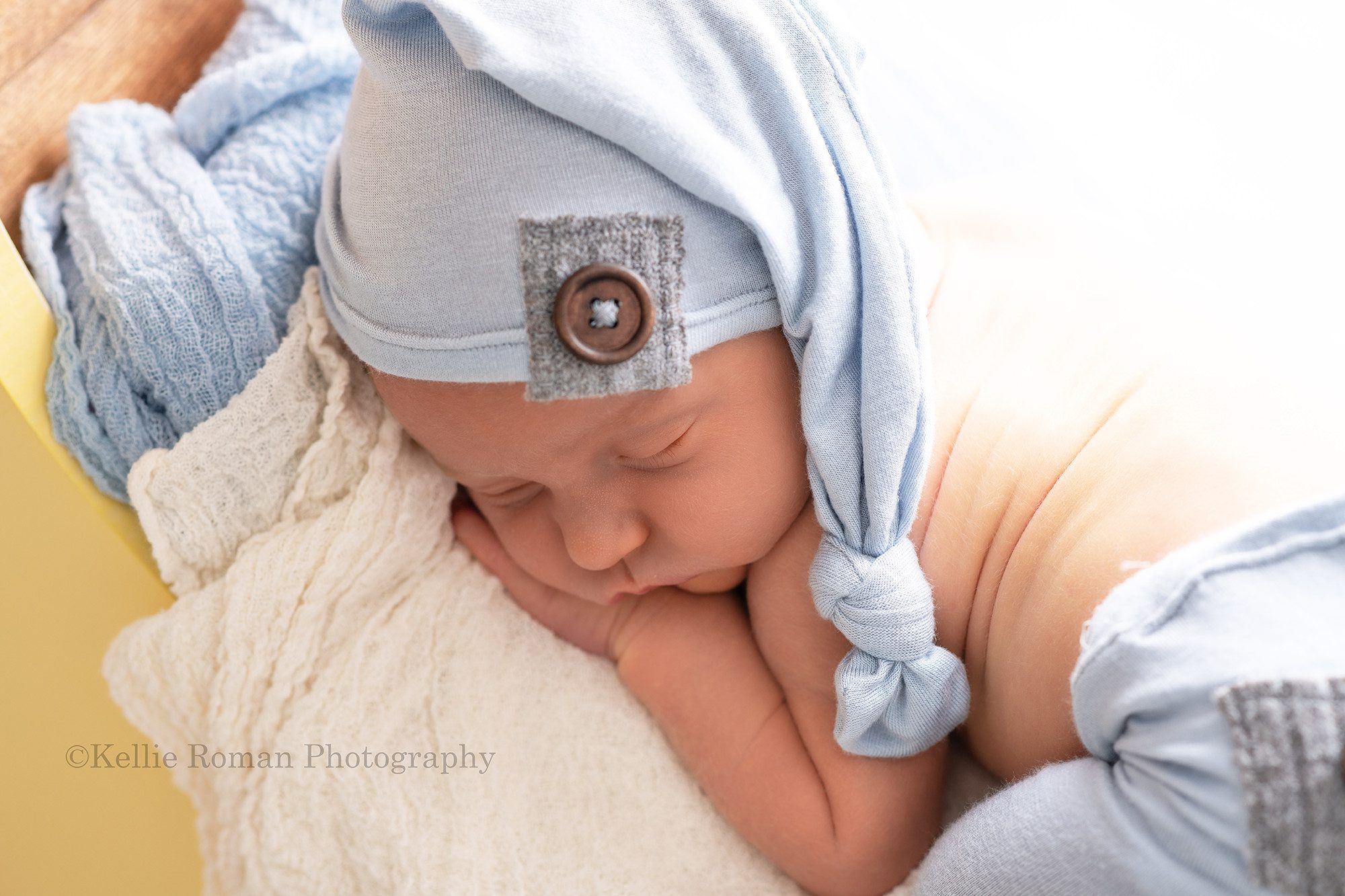 rainbow baby a newborn baby boy posed onto of a yellow wood moon prop in a milwaukee photo studio he's wearing light blue pants and a matching blue sleepy hat he is asleep on his belly with his hands under his child there is backlighting so the image is very bright