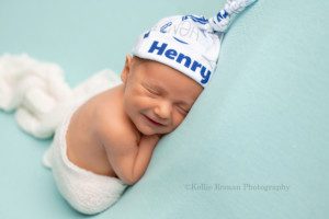 rainbow baby an infant baby boy in a milwaukee photo studio posed onto of a light blue blanket with a white swaddle around his legs he's smiling very big and wearing a white and blue hat with his name Henry on it