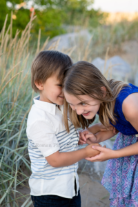popsicle party a brother and sister on a beach in milwaukee Wisconsin holding hands and sharing secrets they are looking at each other and standing in front of tall green beach grass they have on shades of blues and white