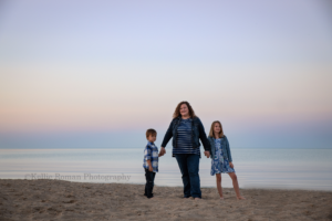 mother and her son and daughter standing on a beach in milwaukee holding hands they are all looking at the camera and the sun has set so the sky has pink and blue pastel shades it's a very simple image