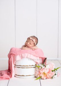 little sister a baby new who is only a few weeks old is posed in a white wood bucket in milwaukee photographers studio the bucket has pink fabric layers in it and pink and white flowers on the side the baby has her chin resting on top of her hands the bucket is on top of a white wood backdrop