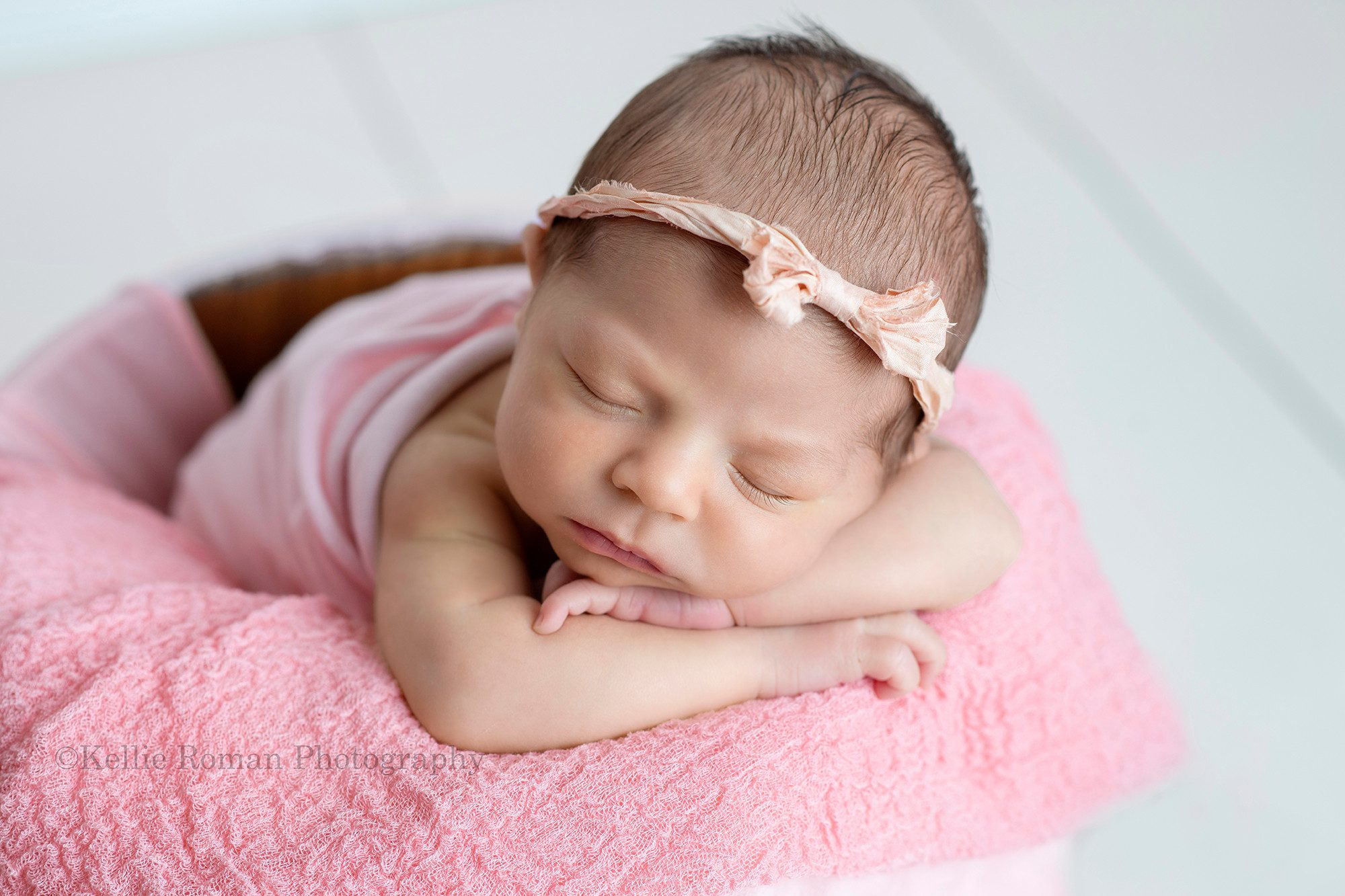 little sister a newborn baby girl is posed in a milwaukee photo studio upright in a white wood bucket there are pink layering fabrics in the bucket the girl has her chin resting on her hands and has a light pink bow headband on