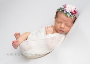little sister a newborn baby girl is posed in milwaukee photo studio on top of a white fabric she is swaddled in a white fabric and laying on her back her two legs are sticking out and her hand is resting on her chest as she sleeps she has on a pink floral headband
