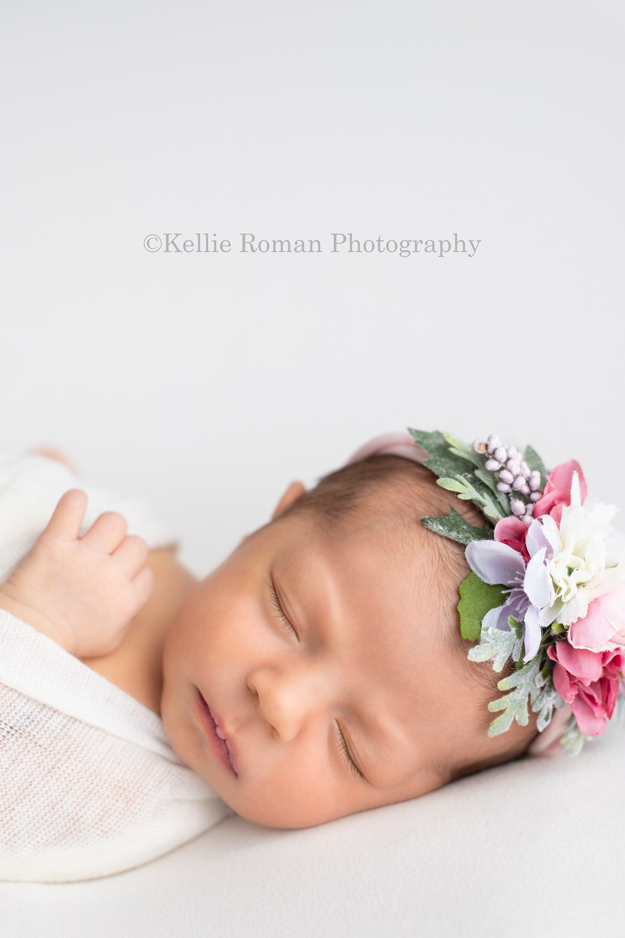 little sister a newborn baby girl in a milwaukee photographers studio is sleeping swaddled in a white wrap she's laying on top of white fabric on her back with one hand up on her chest she has a light pink and purple floral headband on