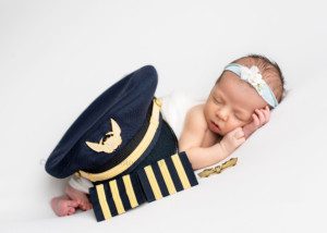 little sister a newborn baby girl in a milwaukee photographer studio posed on white fabric she's sleeping on her side and has her dads pilot hat and wings on top of her