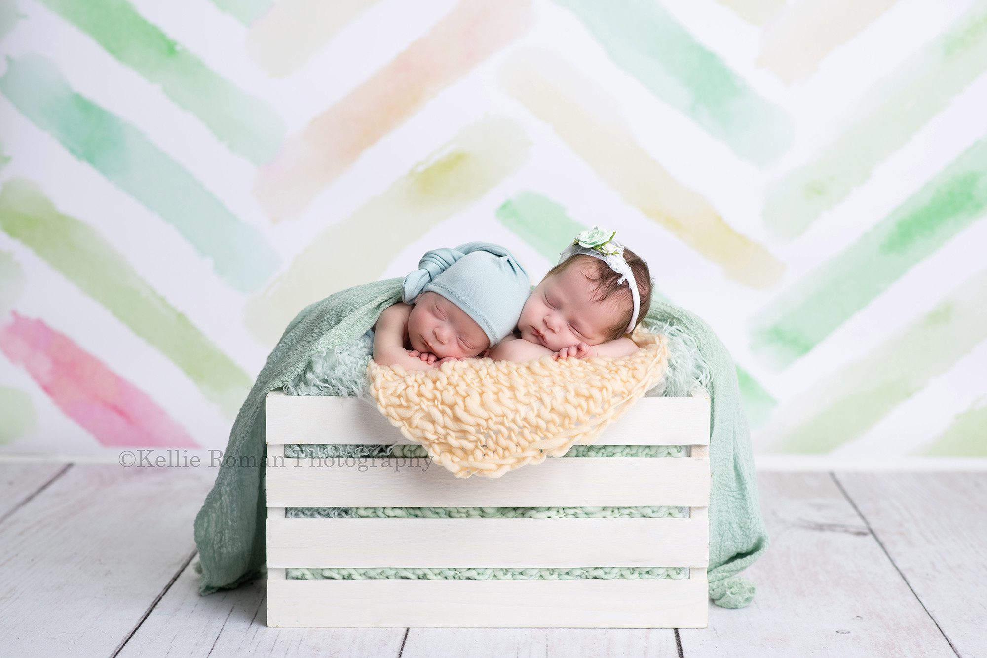 milwaukee twins a brother and newborn twin duo are being photographed in milwaukee Wisconsin photo studio they are posed upright with their chins resting on their arms in a white wood crate the crate is filled with yellow and teal stuffing they are on top of a white wood flooring and a pastel chevron backdrop the babies are both sleeping the girl has a flower headband on and the boy has a blue hat on