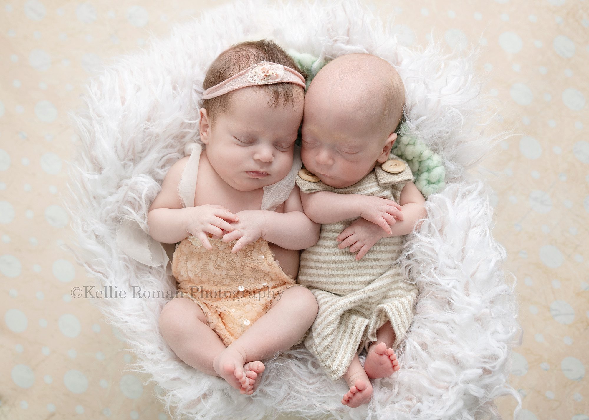 milwaukee twins a newborn brother and sister is posed laying on their backs in a bucket with white fur the boy is wearing a beige striped onesie with buttons and the girl in a peach sequin romper and pink flower headband they are both sleeping with their hands resting on their chests the bucket is on top of a peach polka dot backdrop