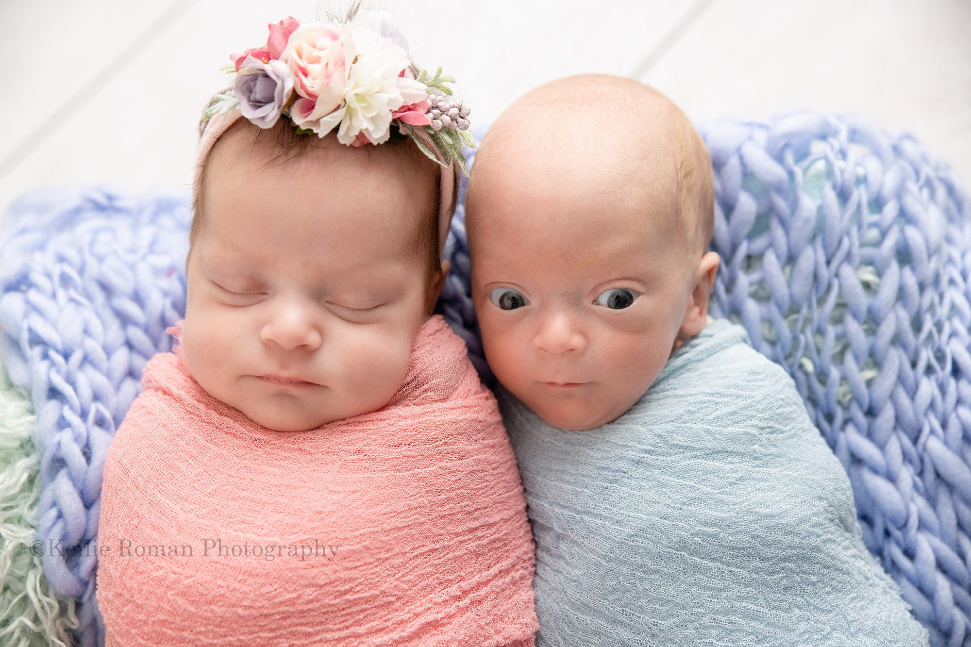 milwaukee twins one boy and one girl newborn twins in milwaukee photo studio they boy has a blue swaddle fabric on and the girl in a pink swaddle fabric the girls has a pink and purple floral headband and the boy is bald they are posed side by side on top of purple knit blanket the boy has his eyes wide open and the girl is sleeping