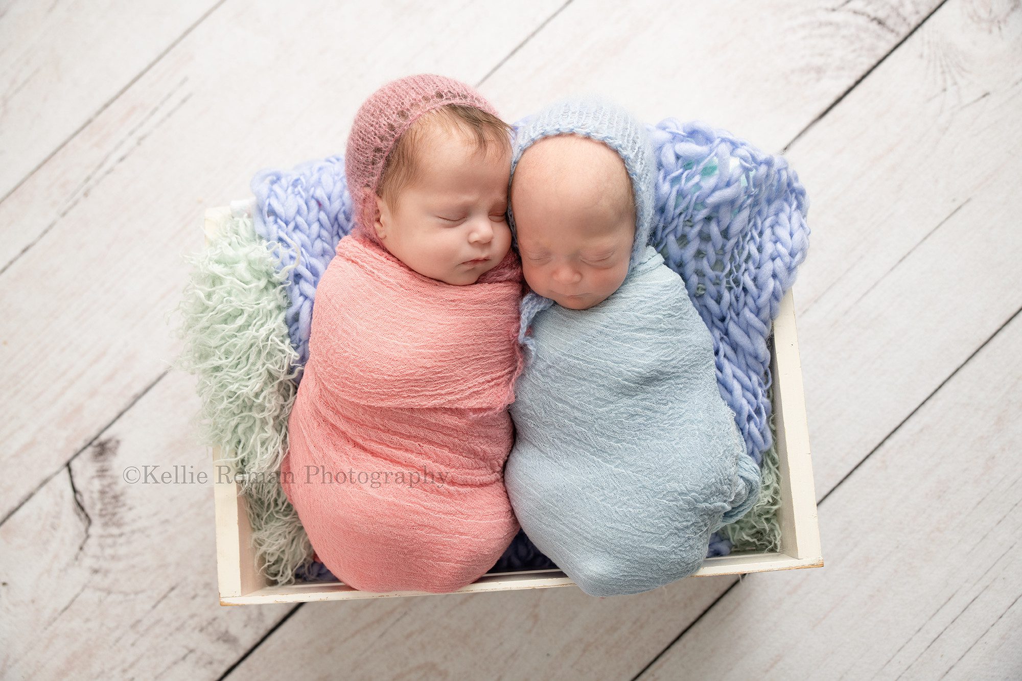 milwaukee twins two newborns one boy and one girl in milwaukee photography studio the girl is wrapped in a pink swaddle with a pink bonnet on the boy is wrapped in a blue swaddle with a blue bonnet on they are sleeping in a white wood crate with teal fur inside of it