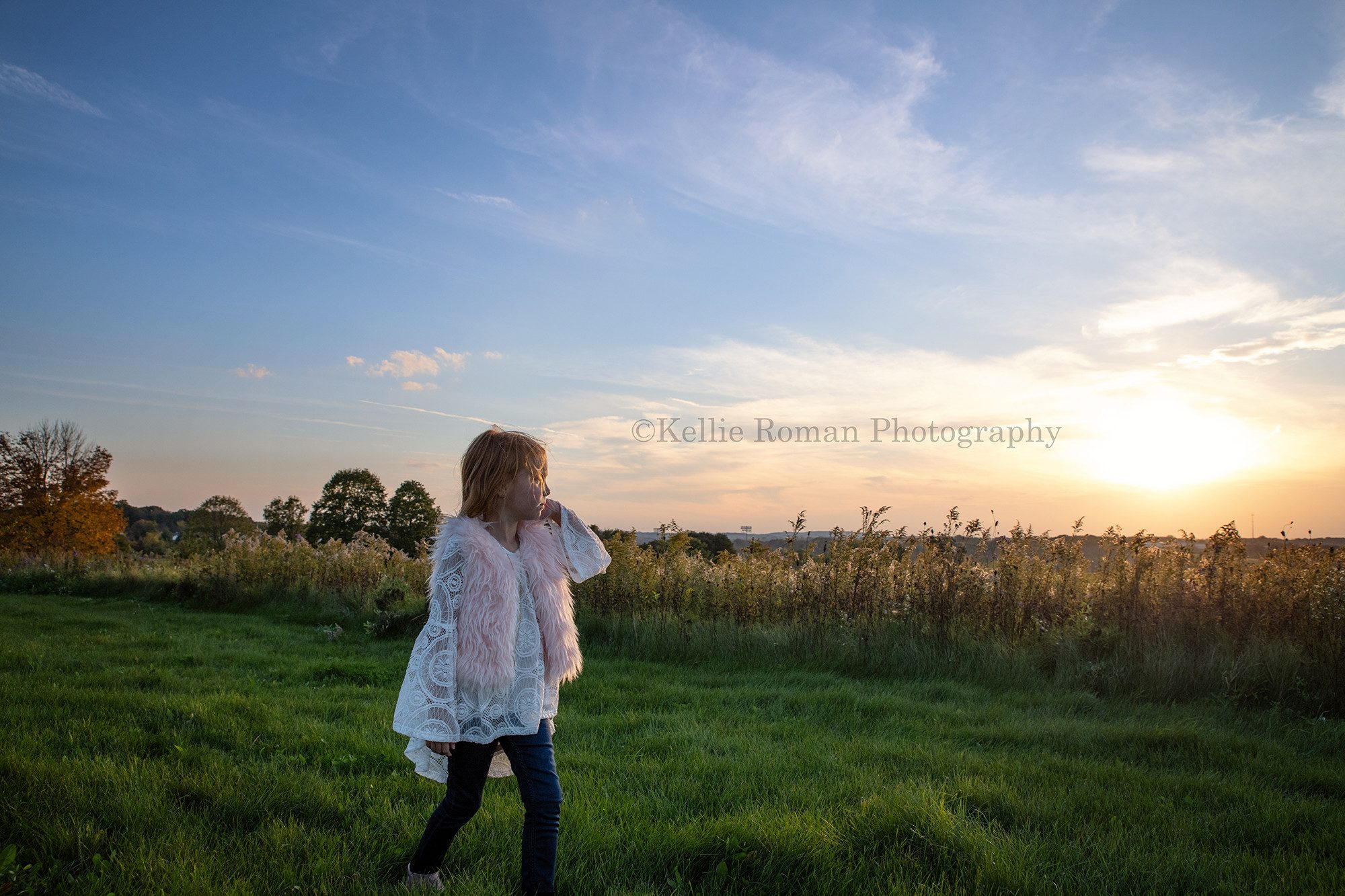 sunset milestone session a young girl wearing jeans a pink fur vest and a crochet white top is walking through green grass in a milwaukee park she is looking off to the side at the horizon which the sun is setting beautiful gold colors