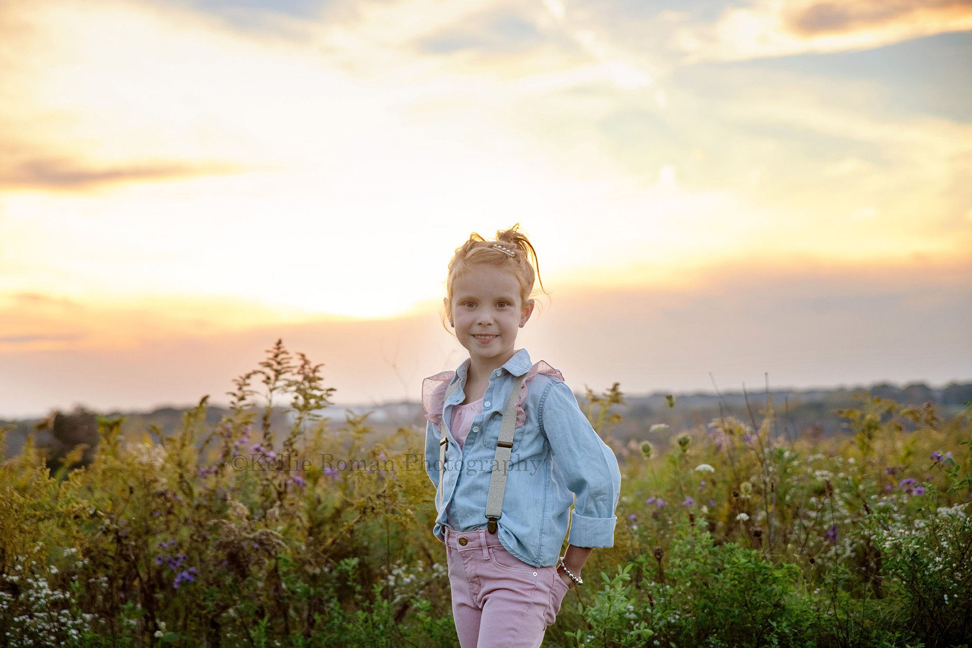 sunset milestone a young girl with red hair is standing in a field of tall grass in milwaukee Wisconsin park she is looking into the camera and smiling the sun is setting behind her on the horizon she has a denim blue shirt with pink and gold suspenders