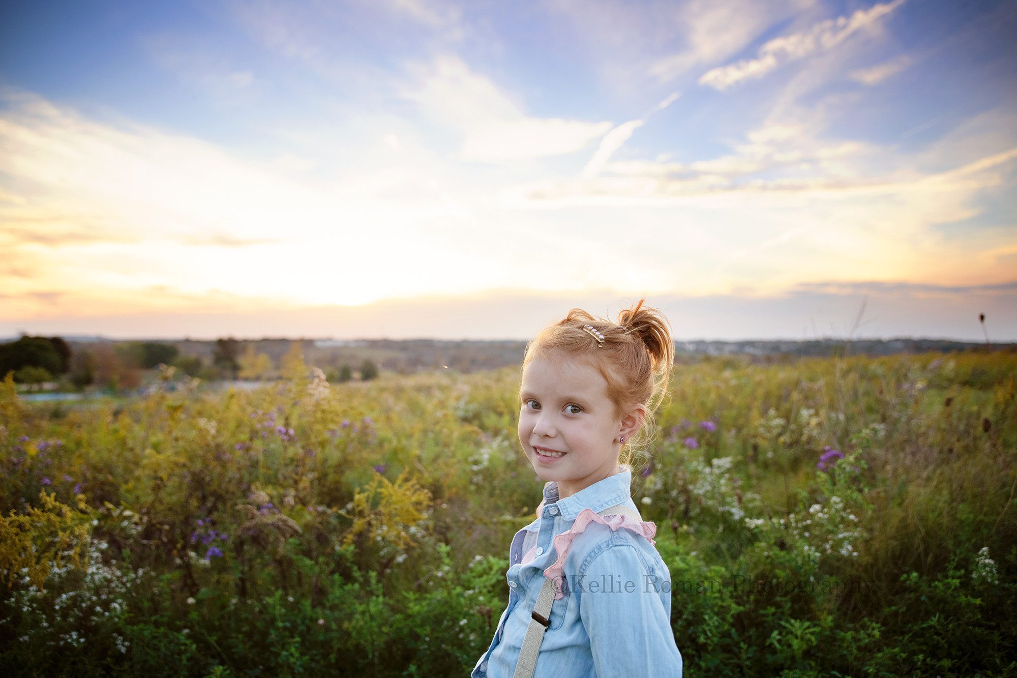 sunset milestone a young girl with red hair is standing in a field of tall grass in milwaukee Wisconsin park she is looking into the camera and smiling the sun is setting behind her on the horizon she has a denim blue shirt with pink and gold suspenders