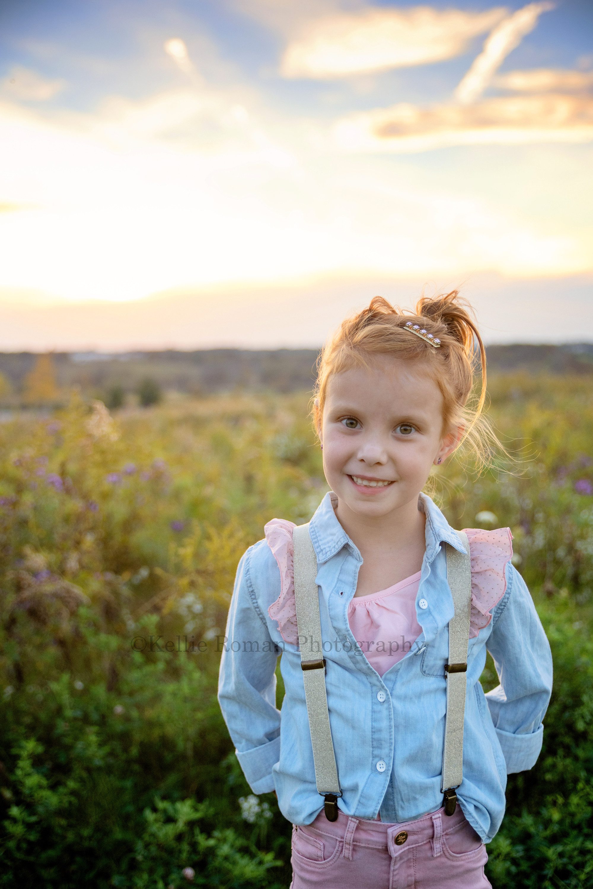 sunset milestone a young girl in milwaukee county Wisconsin is in a field of tall green grass with a bright colorful sunset behind her she's wearing pink jeans with a pink take top under a blue denim shirt she has on pink and gold suspenders she has red hair and it's in a messy pony tail