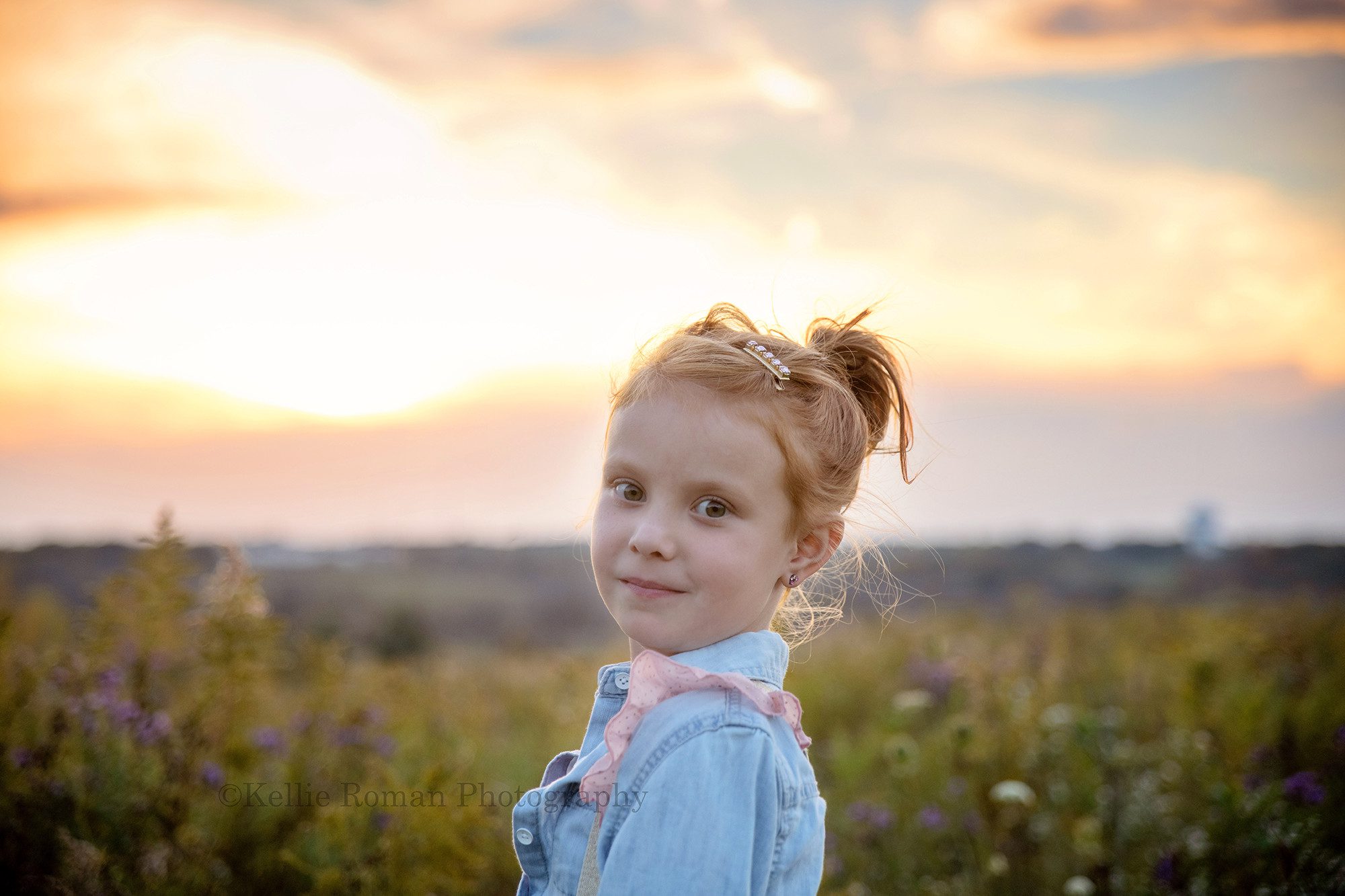 sunset milestone a young girl in milwaukee standing in a field of tall grass the with sun setting on the horizon behind her the sky is beautiful shades of blue pink and orange the girl has red hair and it's up in a messy bun she is wearing a denim shirt and pink and gold suspenders