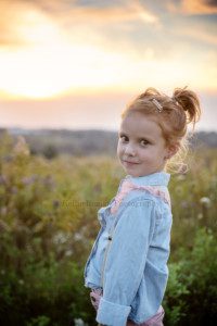sunset milestone a six year old. girl with red hair is standing in a field of green grass in milwaukee the horizon is the sunset which are shades of pink and orange the girl is wearing a blue denim shirt with pink and gold suspenders and is looking into the camera