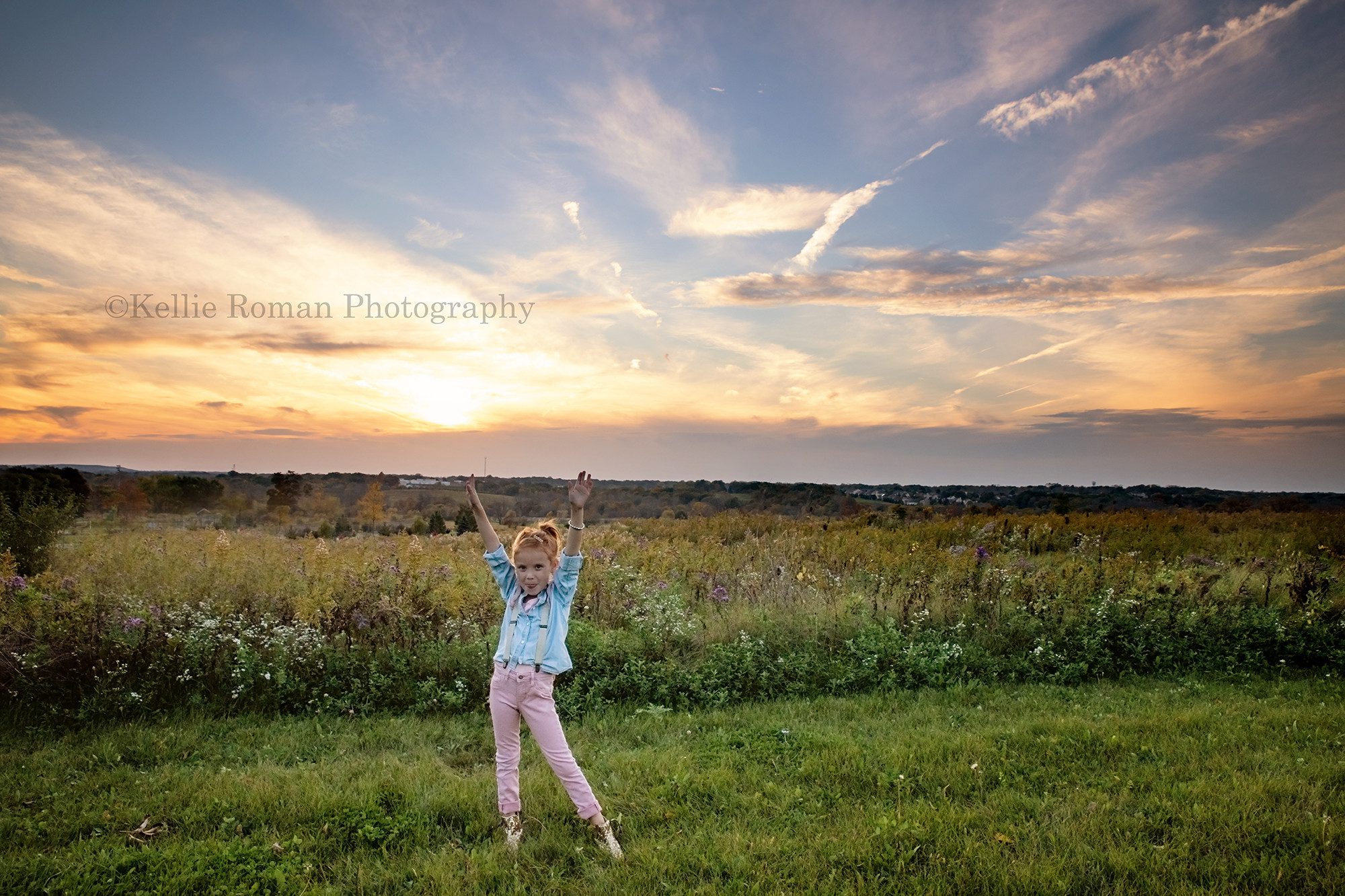young girl standing in a field of tall green grass in milwaukee doing her six year sunset milestone session the sun is setting behind her and the sky is gorgeous shades of oranges blues and pinks the girl is wearing pink pants with a denim shirt and suspenders that are gold and pink she's holding her hands up in the air
