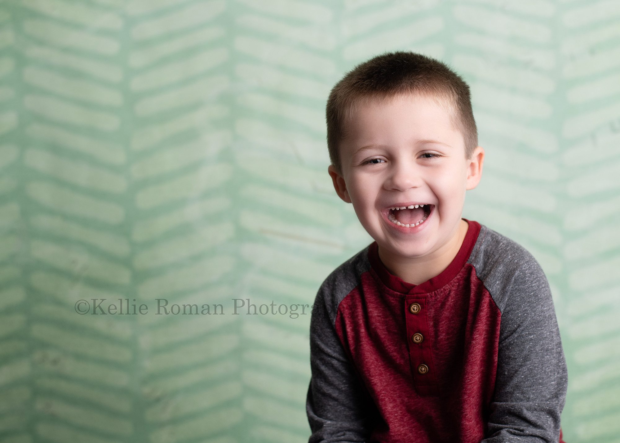 milwaukee milestone session a four year old boy in milwaukee photographers studio is smiling really big into the camera he's in front of a green chevron backdrop and is wearing a red and grey shirt