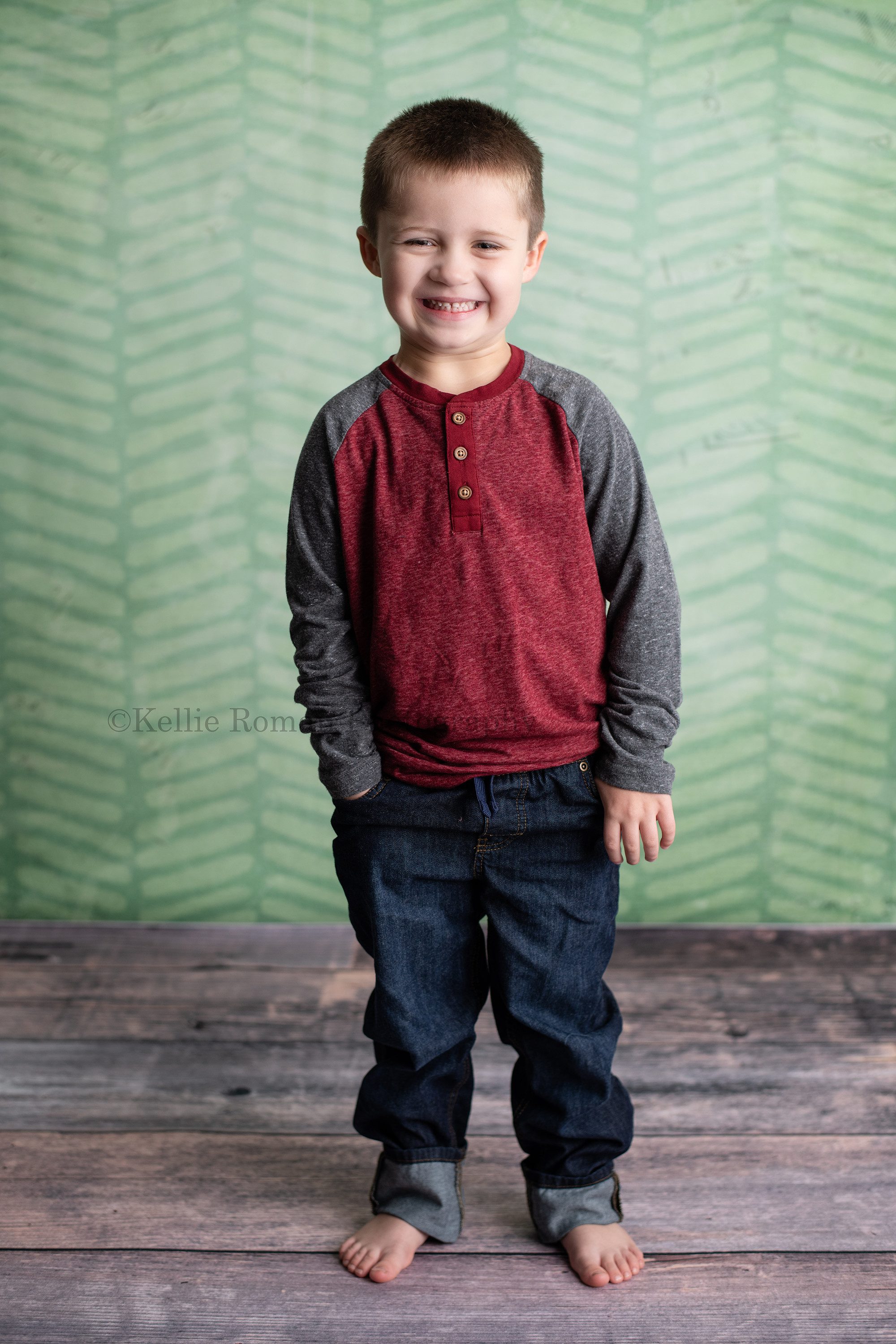 milwaukee milestone photographer a young boy in milwaukee photographers studio standing on dark wood floor with a green chevron backdrop he's wearing jeans that are cuffed and red and grey shirt he has his right hand in his pocket and is smiling really big
