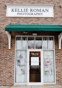 greendale village studio a photographers store front in the historic downtown greendale village the store front has six large silk banners in the front windows and a vinyl logo on the door a large sign of kellie roman photograph is above the doorway the building is all red brick