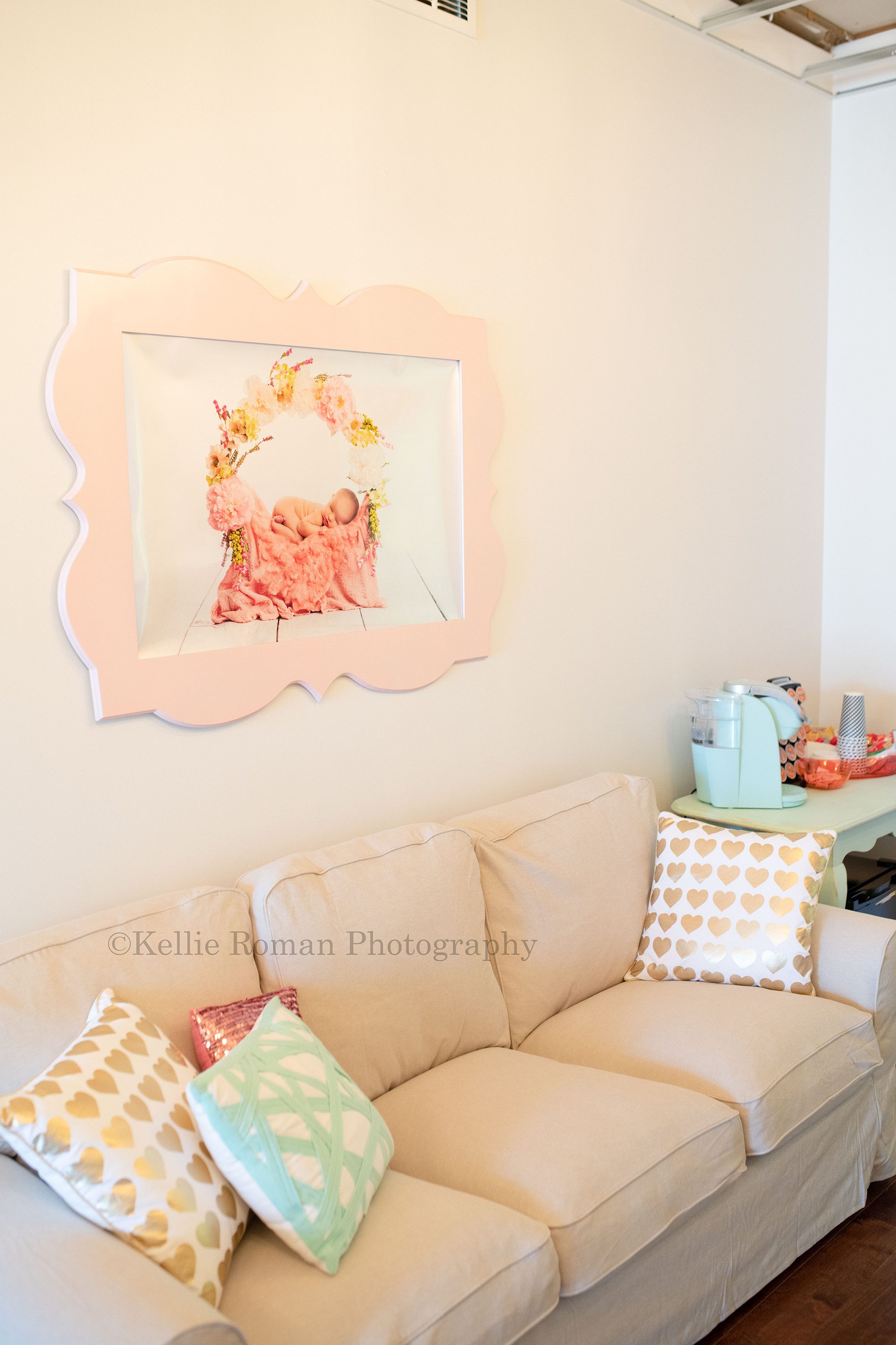 greendale village studio a photographer studio space located in the historic downtown greendale village in milwaukee county the image is a parent lounge with ivory seating area and pastel and gold pillows on top a pink large frame on the wall with a newborn baby photo inside a coffee and snack table is off to the side