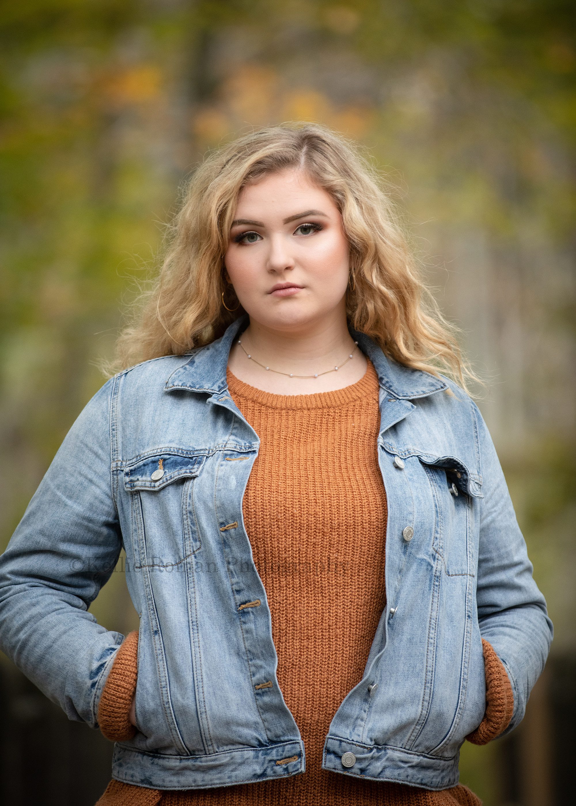milwaukee senior photographer a young girl looking into the camera with a senior face she is blonde curly hair and blue eyes she's wearing a jean jacket with an orange ribbed sweater underneath she has both hands in her pockets