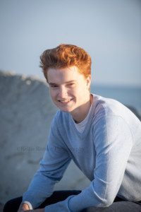 milwaukee photographer a high school senior boy is sitting on a large rock at lake Michigan on the beach in kenosha Wisconsin he's wearing a light blue sweater and jeans