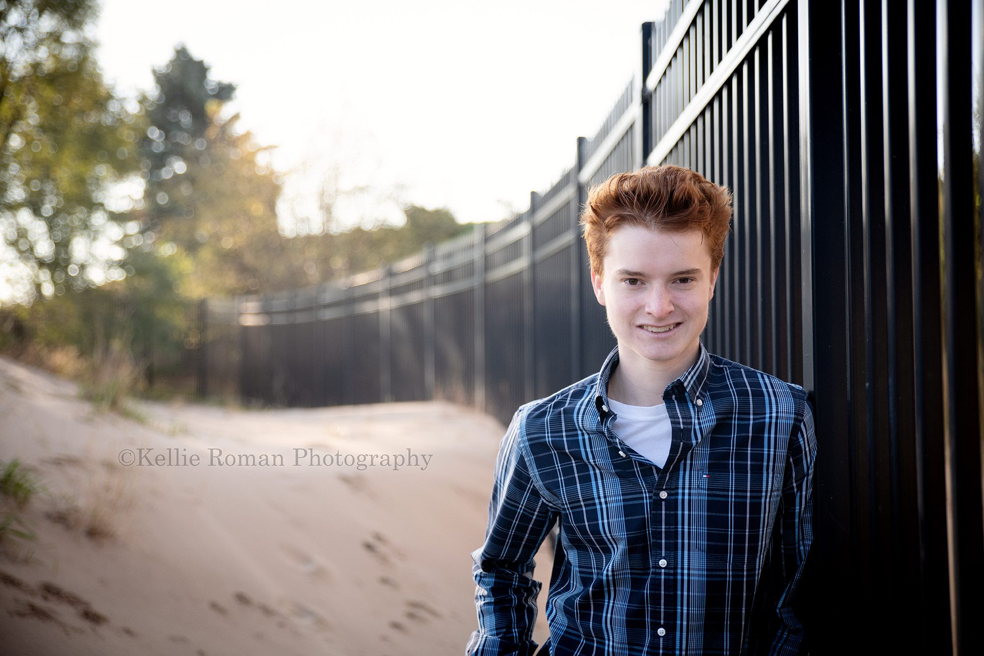 milwaukee photographer a high school senior boy is standing next to a rod iron black fence on the beach in kenosha Wisconsin there is stand behind him and he is wearing a black and blue plaid shirt he has red hair and is looking into the camera