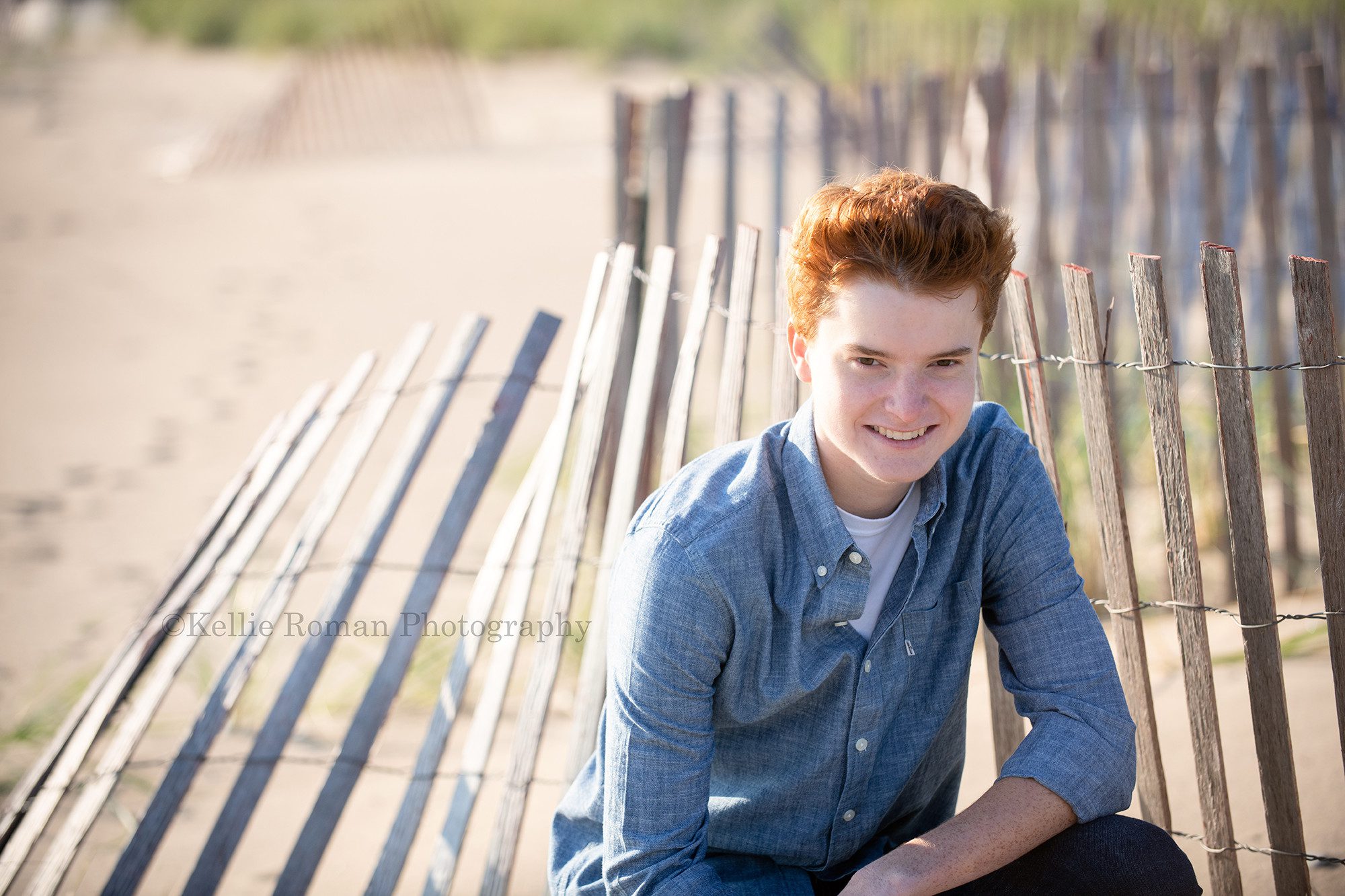 milwaukee photographer a high school senior guy is being photographed on kenosha Wisconsin beach he's kneeling in front of a rustic beach fence and is wearing a denim button up shirt he has red hair and is smiling into the camera
