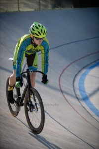 milwaukee photographer a high school senior guy is riding his bike at the kenosha velodrome its an action shot of him riding he has a helmet and gloves on as well as sunglasses and riding shoes his cycling uniform is green and blue the sun is shining from behind him on the race track