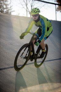 milwaukee photographer a high school senior boy is riding his bike at the kenosha velodrome he's wearing his cycling uniform which is neon green and blue he has a helmet gloves and riding shoes on with sunglasses the image is with the sun shining behind him