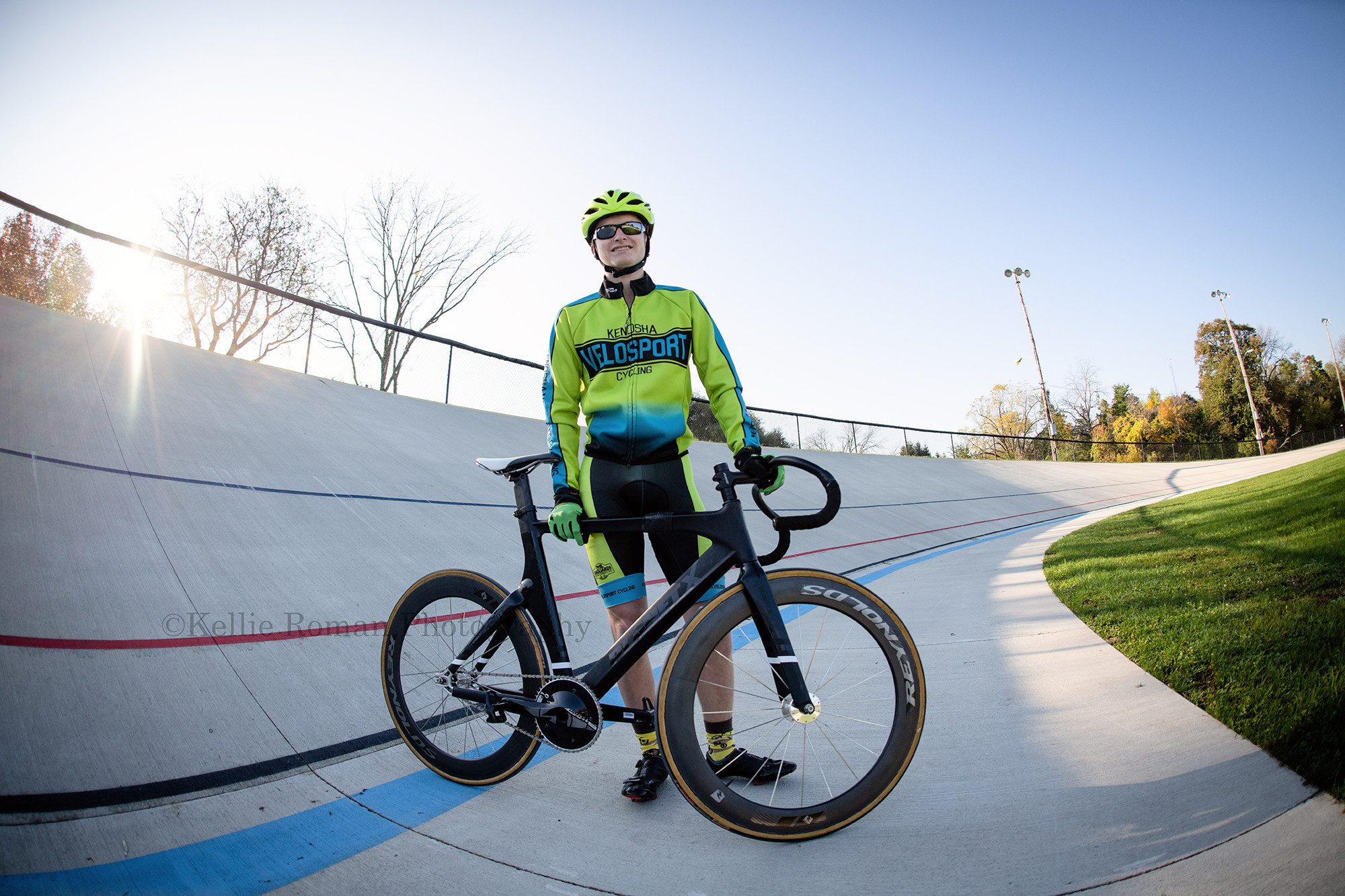 milwaukee photographer a high school senior guy is at the kenosha velodrome with his cycling uniform on he's standing behind his bike on the cycling track he's looking at the camera and smiling the image is taken at a very wide angle