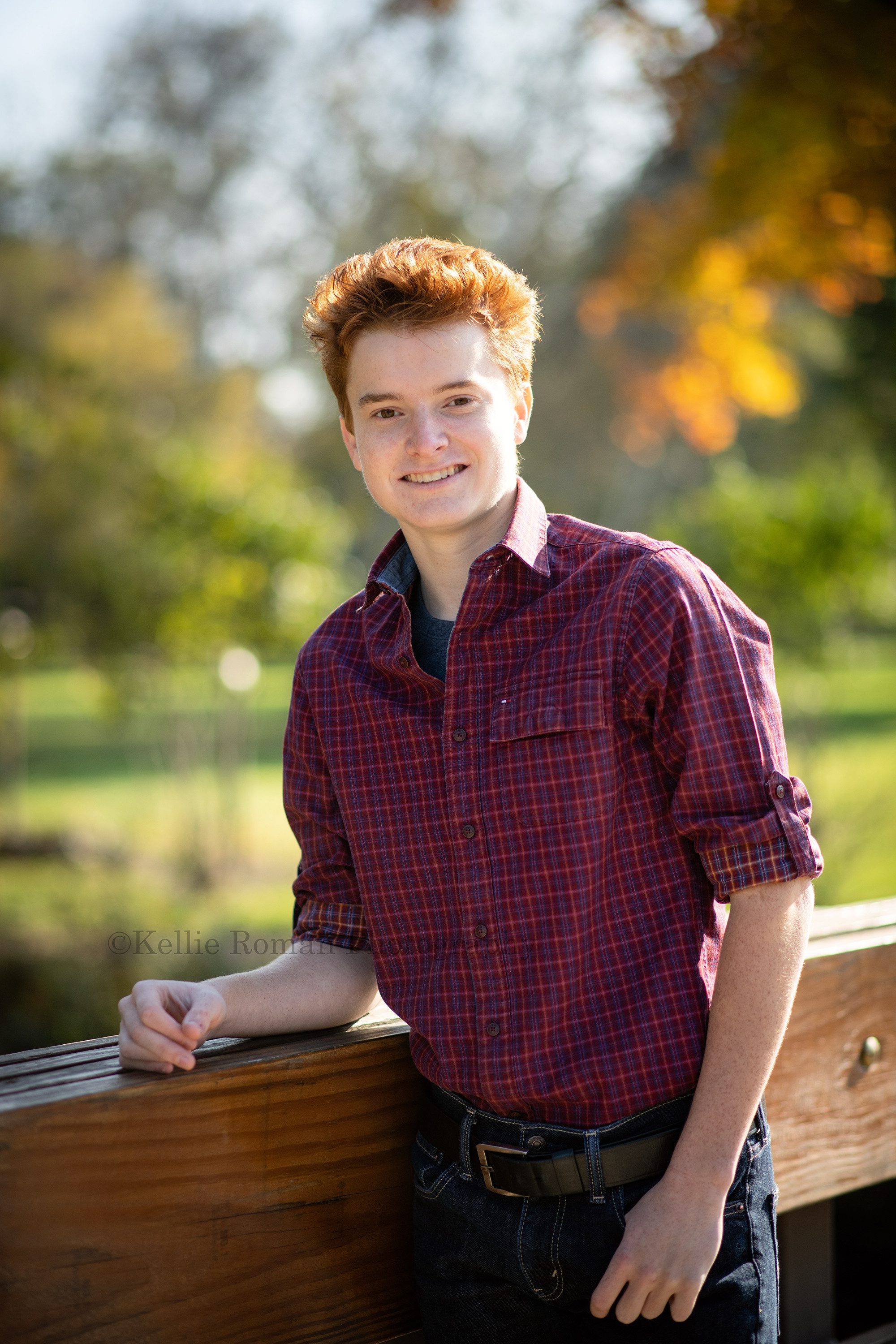milwaukee photographer a young high school senior boy standing leaning against a wood bridge at a kenosha park he has on jeans and a red checkered shirt he has his one thumb in his pocket and the other arm resting on the bridge he's smiling at the camera and has red hair